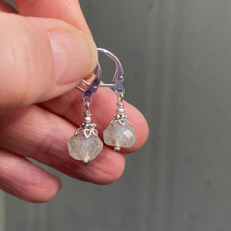 May include: A pair of silver leverback earrings. Each earring features a faceted, pale green gemstone pendant with a decorative silver cap and a small silver bead at the bottom. The earrings are held up by a person's fingers.