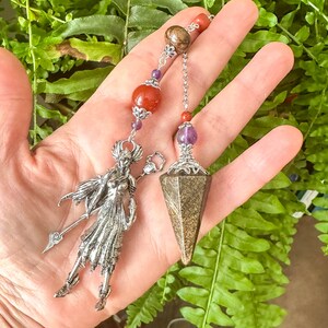 May include: A silver pendulum featuring a woman figure and a faceted, brown crystal point. The pendulum is decorated with red and purple beads, and a silver chain. The background is green foliage.