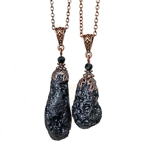 May include: Two black pendant necklaces with textured, teardrop-shaped stones. The pendants are suspended from copper-colored chains and feature ornate copper-colored caps and black beads. The background is white.