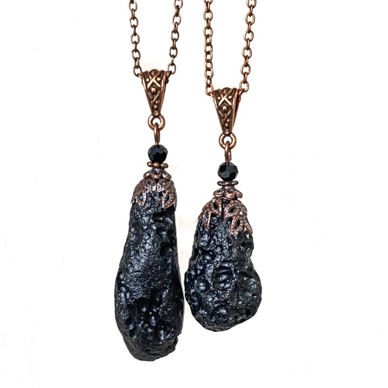 May include: Two black pendant necklaces with textured, teardrop-shaped stones. The pendants are suspended from copper-colored chains and feature ornate copper-colored caps and black beads. The background is white.