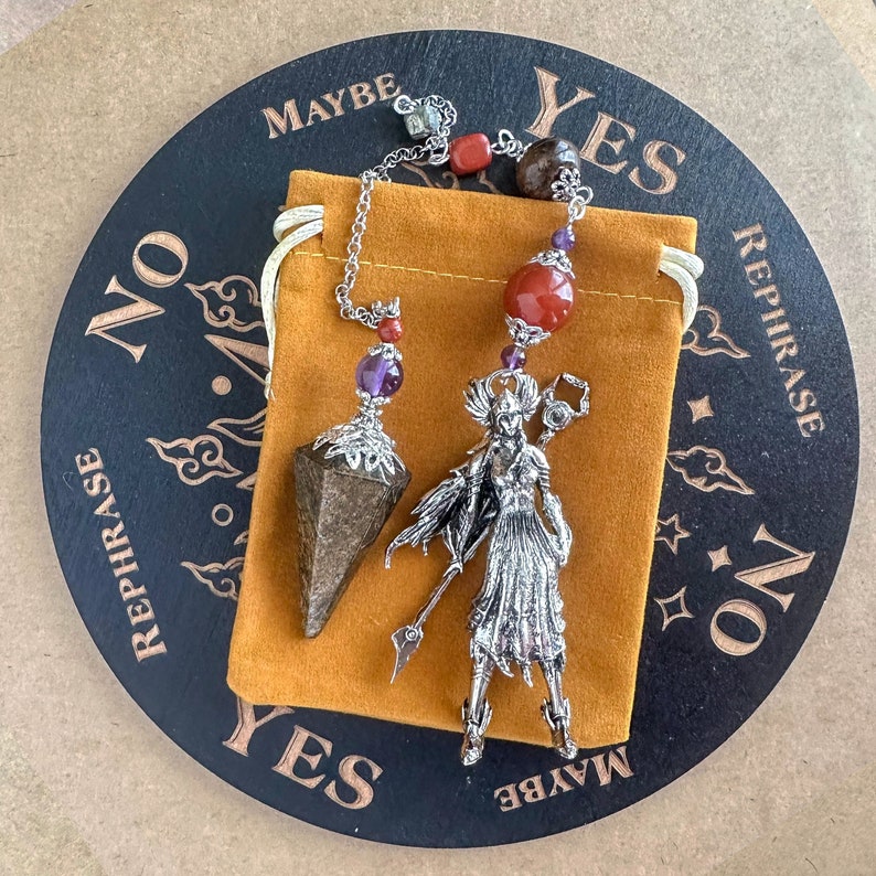 May include: A silver pendulum and a warrior figurine with a spear, decorated with red and purple beads, sit on a mustard-coloured pouch. The pendulum features a faceted, brown crystal. A round, black board with the words "YES", "NO", "MAYBE", and "REPHRASE" surrounds the objects.