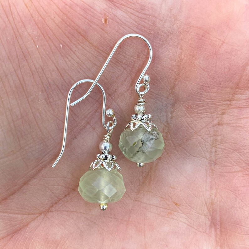 May include: A pair of silver dangle earrings featuring faceted, pale green gemstone beads. The earrings have a decorative silver cap and a simple hook closure. The gemstones have a unique, textured appearance, adding to their appeal.