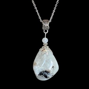 May include: A silver chain necklace with a large, polished, teardrop-shaped moonstone pendant. The moonstone is primarily white with black and gray inclusions. The pendant is accented with silver-colored metalwork and a small, round bead.