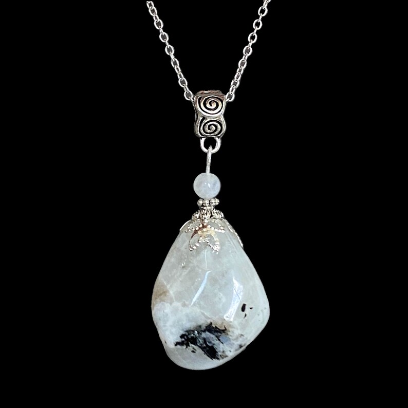 May include: A silver chain necklace with a large, polished, teardrop-shaped moonstone pendant. The moonstone is primarily white with black and gray inclusions. The pendant is accented with silver-colored metalwork and a small, round bead.