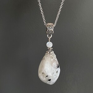 May include: A silver necklace featuring a large, teardrop-shaped, white and gray stone pendant. The pendant is accented with black inclusions and a decorative silver cap. The necklace chain is delicate, and the design includes a small, round, white stone.