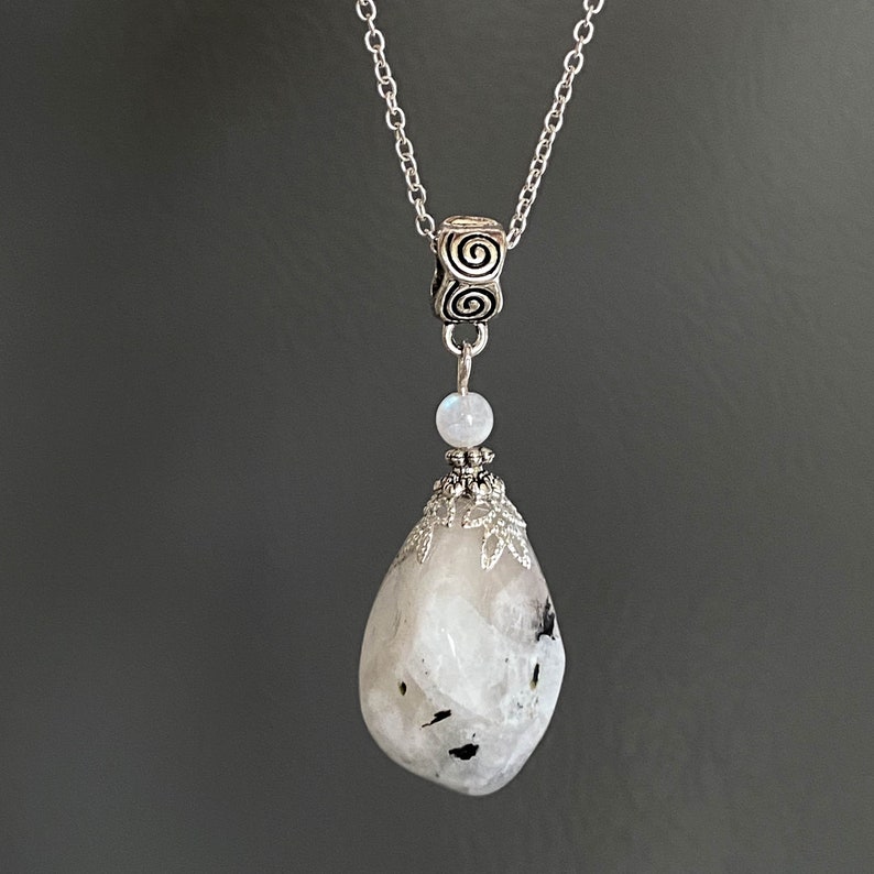 May include: A silver necklace featuring a large, teardrop-shaped, white and gray stone pendant. The pendant is accented with black inclusions and a decorative silver cap. The necklace chain is delicate, and the design includes a small, round, white stone.