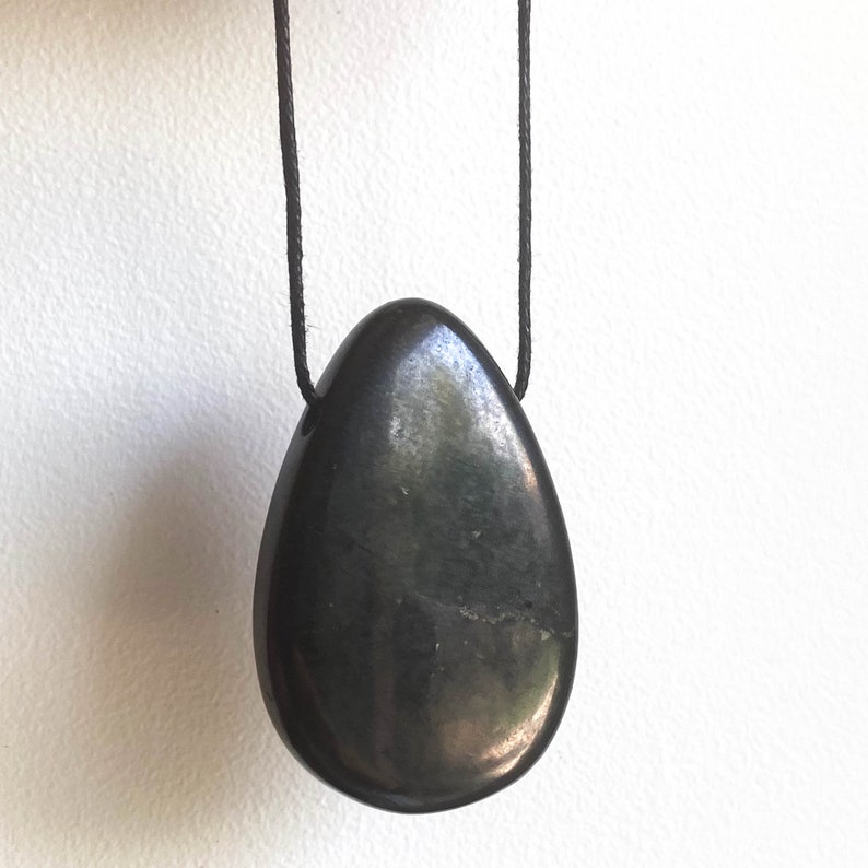May include: A dark gray, teardrop-shaped pendant hangs from a black cord. The polished stone has a smooth, reflective surface. The pendant is suspended against a plain white background. The jewelry is a necklace.