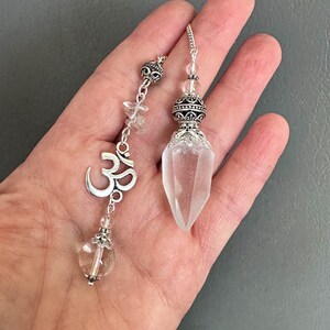 K&ouml;nnte beinhalten: Zwei Kristallpendel mit silberfarbenen Metallakzenten. Eines zeigt ein Om-Symbol und eine klare Kristallkugel. Das andere hat eine facettierte Kristallspitze. Beide sind an Ketten mit dekorativen Perlen aufgeh&auml;ngt.