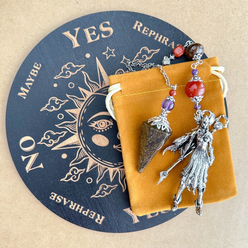 May include: A divination set featuring a black Ouija board with gold lettering and a sun design. A brown crystal pendulum with silver accents and a silver figurine are displayed with an orange pouch. The board reads "YES", "NO", "MAYBE", and "REPHRASE".