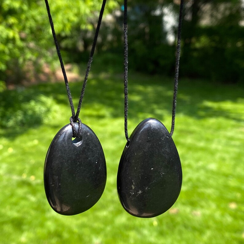 May include: Two black, teardrop-shaped pendants hanging from black cords. The pendants have a smooth, polished surface and are displayed against a blurred green background. The pendants are likely made of stone or a similar material.