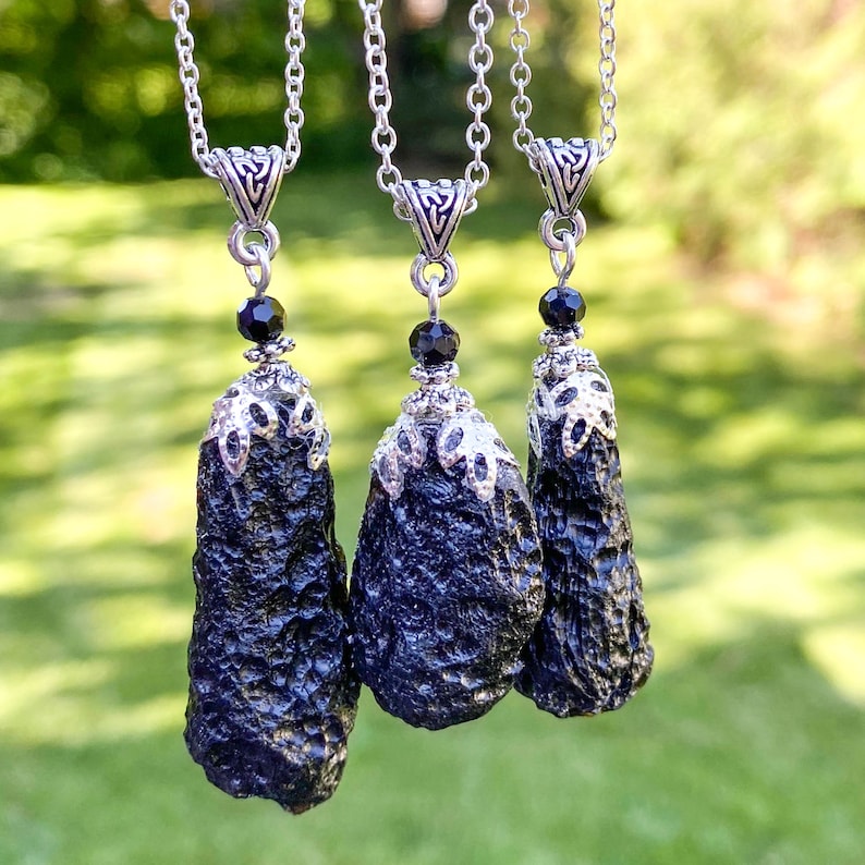 May include: Three black tektite pendants with silver-colored metal accents and chains. Each pendant has a unique, textured surface and is suspended from a delicate chain. A small black bead is placed above the pendant.