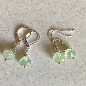 May include: A pair of earrings with faceted, pale green gemstones. The earrings have silver-colored lever-back closures and decorative silver accents. The gemstones are a soft, translucent green color and are cut into a rounded shape.