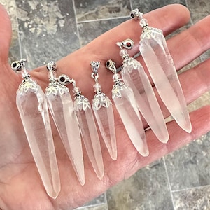 May include: A collection of clear quartz crystal pendants with silver-colored metal caps and decorative accents. The pendants vary slightly in size and are displayed against a neutral background.