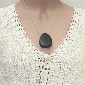 May include: A dark gray, teardrop-shaped pendant necklace. The pendant hangs from a delicate silver chain. The necklace is displayed against a cream-colored, crocheted top.