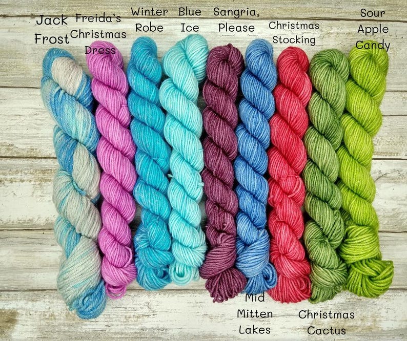 May include: Nine skeins of yarn in various colors, including blue, green, pink, and purple. The yarn is arranged vertically and labeled with names like "Jack Frost", "Freida's Christmas Dress", "Winter Robe", "Blue Ice", "Sangria, Please", "Christmas Stocking", "Sour Apple Candy", "Mid Mitten Lakes", and "Christmas Cactus".