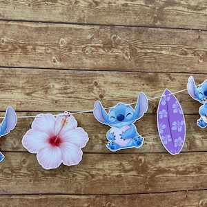 Op de afbeelding: Een slinger met blauwe cartoonfiguren, een roze hibiscusbloem en een paars surfplank. De figuren houden een hart vast en spelen ukulele. De slinger is geregen aan een witte draad tegen een houten achtergrond.