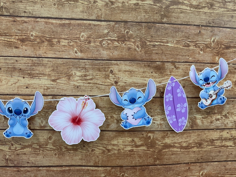 Op de afbeelding: Een slinger met blauwe cartoonfiguren, een roze hibiscusbloem en een paars surfplank. De figuren houden een hart vast en spelen ukulele. De slinger is geregen aan een witte draad tegen een houten achtergrond.