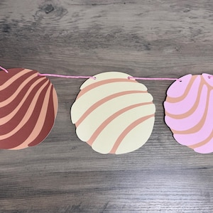 May include: A decorative banner featuring paper cutouts shaped like conchas, a traditional Mexican sweet bread. The conchas are in shades of brown, cream, and pink, strung together with pink twine. The banner is displayed against a wooden background.
