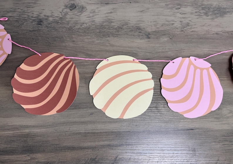 May include: A decorative garland featuring circular paper cutouts shaped like concha sweet bread rolls. The cutouts are in shades of brown, cream, and light pink, each displaying a textured shell-like pattern. They are strung together on a thin pink string.