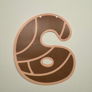 May include: A flat, die-cut paper decoration in the shape of the number six. The design features a dark brown, textured surface divided into segments by light peach-colored outlines. Two small circular holes are punched at the top for hanging.