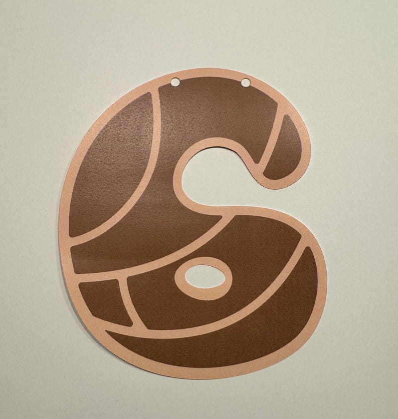 May include: A flat, die-cut paper decoration in the shape of the number six. The design features a dark brown, textured surface divided into segments by light peach-colored outlines. Two small circular holes are punched at the top for hanging.