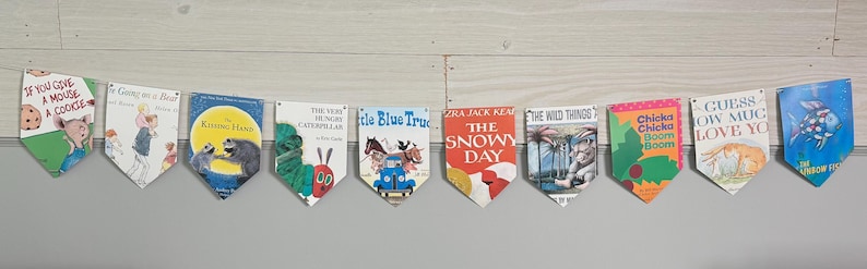 Puede incluir: Una guirnalda de portadas de libros infantiles, incluyendo "The Very Hungry Caterpillar" y "The Snowy Day". Los banderines son de varios colores y presentan ilustraciones de los libros. La guirnalda cuelga sobre un fondo blanco y gris.