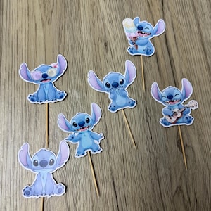 Op de afbeelding: Set van zeven blauwe Stitch-cupcaketoppers. Elke topper toont een andere pose van het personage, sommige met accessoires zoals een zonnebril, ijs en een ukelele. De toppers zijn bevestigd aan houten stokjes.