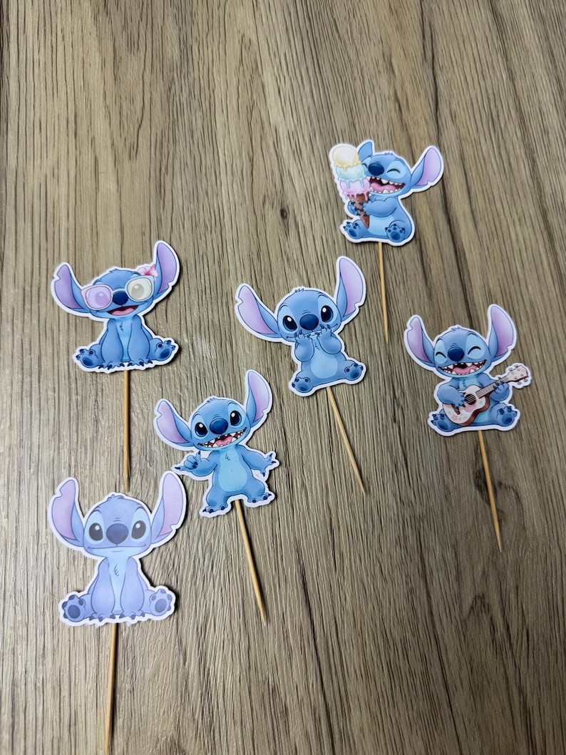 Op de afbeelding: Set van zeven blauwe Stitch-cupcaketoppers. Elke topper toont een andere pose van het personage, sommige met accessoires zoals een zonnebril, ijs en een ukelele. De toppers zijn bevestigd aan houten stokjes.
