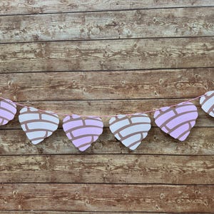 May include: A decorative garland featuring six heart-shaped cutouts designed to resemble concha sweet bread. Each heart has a light pink base with a white, textured, shell-like pattern on the front, all strung together on a thin pink string.