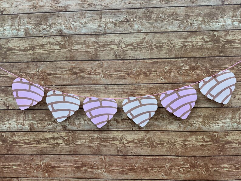 May include: A decorative garland featuring six heart-shaped cutouts designed to resemble concha sweet bread. Each heart has a light pink base with a white, textured, shell-like pattern on the front, all strung together on a thin pink string.