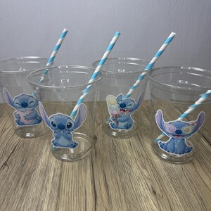 Op de afbeelding: Transparante plastic bekers met Stitch-personage-stickers. Elke beker heeft een blauw-wit gestreept rietje. De bekers zijn gerangschikt op een houten oppervlak.