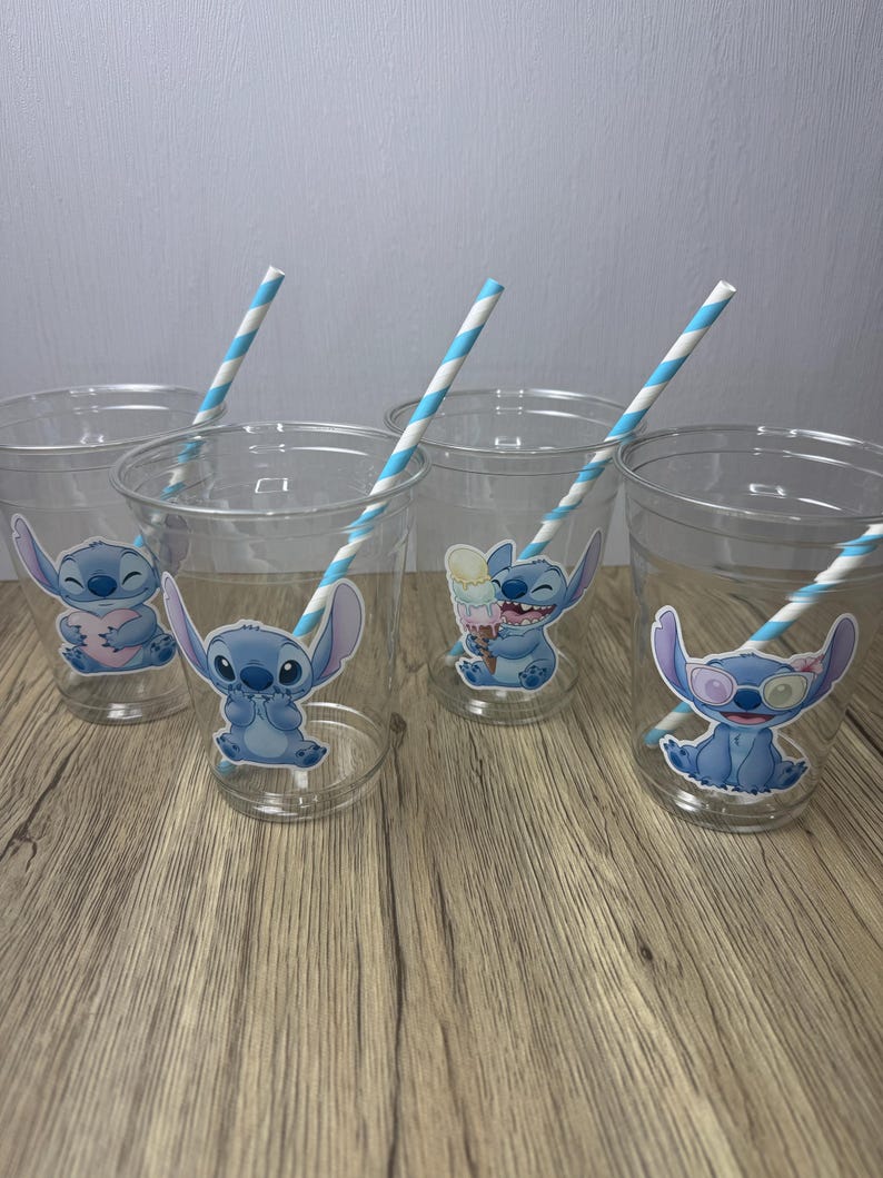 Op de afbeelding: Transparante plastic bekers met Stitch-personage-stickers. Elke beker heeft een blauw-wit gestreept rietje. De bekers zijn gerangschikt op een houten oppervlak.