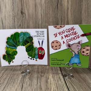 Puede incluir: Se muestran dos portadas de libros infantiles. Una es "The Very Hungry Caterpillar" con una colorida ilustraci&oacute;n de oruga. La otra es "If You Give a Mouse a Cookie", que muestra un rat&oacute;n con galletas. Ambas se sujetan con soportes de alambre plateado.