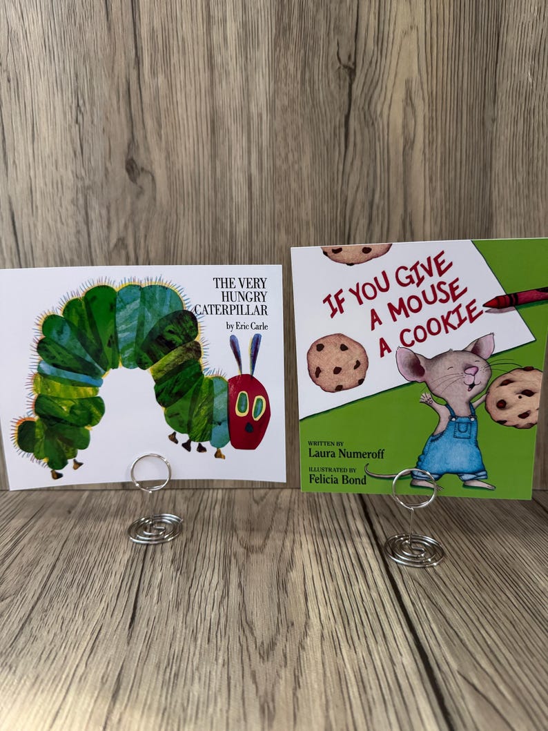 Puede incluir: Se muestran dos portadas de libros infantiles. Una es "The Very Hungry Caterpillar" con una colorida ilustraci&oacute;n de oruga. La otra es "If You Give a Mouse a Cookie", que muestra un rat&oacute;n con galletas. Ambas se sujetan con soportes de alambre plateado.
