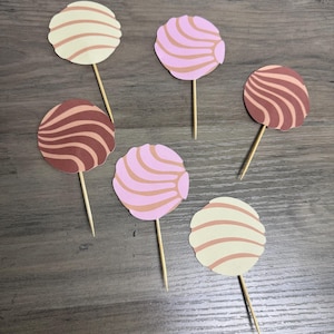 May include: Six circular paper cupcake toppers shaped like concha sweet breads, each attached to a wooden toothpick. The toppers feature swirl patterns in cream, light pink, and dark brown colors, arranged in a scattered layout on a flat surface.