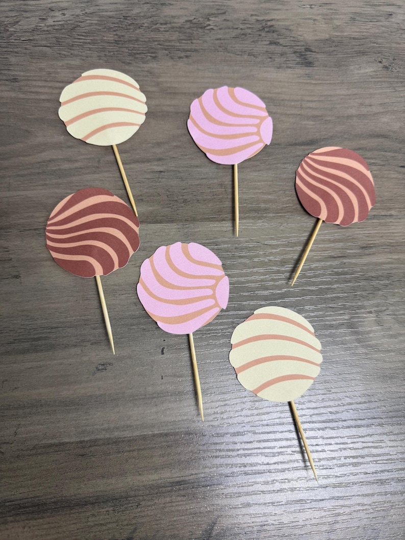 May include: Six circular paper cupcake toppers shaped like concha sweet breads, each attached to a wooden toothpick. The toppers feature swirl patterns in cream, light pink, and dark brown colors, arranged in a scattered layout on a flat surface.