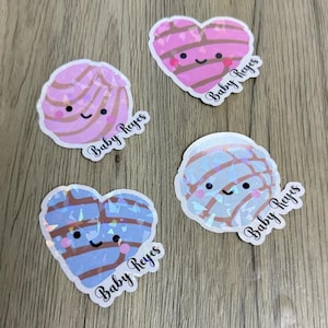 May include: Four stickers featuring cute, smiling concha bread designs. Two are round and two are heart-shaped, with pink and blue colors and a holographic finish. Each sticker includes the text Baby Reyes in a black cursive script font.