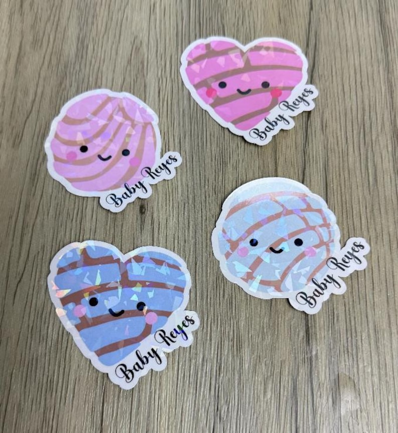 May include: Four stickers featuring cute, smiling concha bread designs. Two are round and two are heart-shaped, with pink and blue colors and a holographic finish. Each sticker includes the text Baby Reyes in a black cursive script font.