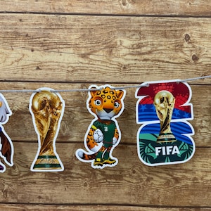 May include: A decorative banner featuring cutouts of a cartoon eagle mascot, the FIFA World Cup trophy, a cartoon leopard mascot holding a soccer ball, and a graphic with the text 2022 FIFA, all strung together on a thin silver cord.