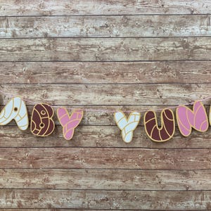 May include: A decorative banner spelling out BABY YUNA, featuring individual letter cutouts designed to resemble concha sweet bread in shades of pink, white, and brown, all strung together on a thin light pink string for hanging display.