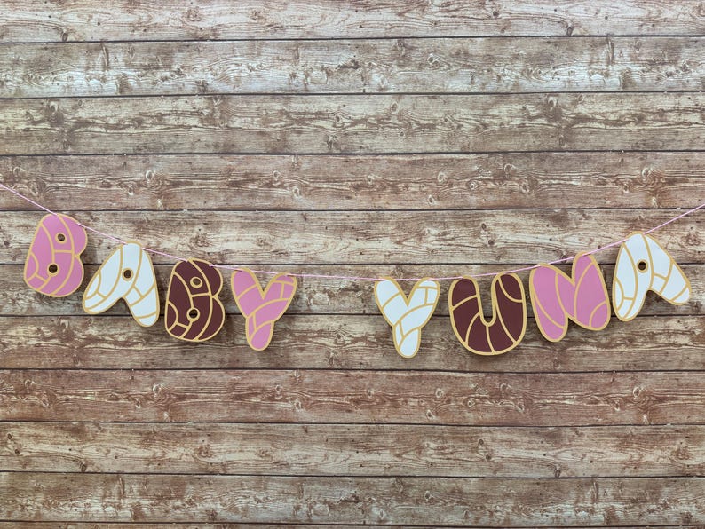 May include: A decorative banner spelling out BABY YUNA, featuring individual letter cutouts designed to resemble concha sweet bread in shades of pink, white, and brown, all strung together on a thin light pink string for hanging display.
