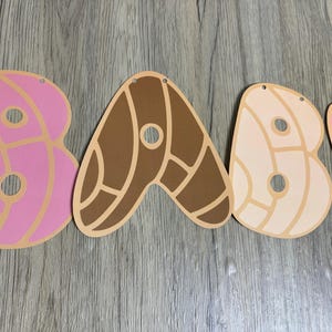 May include: A decorative banner spelling the word BABY with each letter shaped like a concha sweet bread. The letters are pink, dark brown, and light beige, featuring a distinct shell-like pattern with circular cutouts and small holes for hanging.