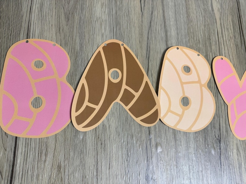 May include: A decorative banner spelling the word BABY with each letter shaped like a concha sweet bread. The letters are pink, dark brown, and light beige, featuring a distinct shell-like pattern with circular cutouts and small holes for hanging.