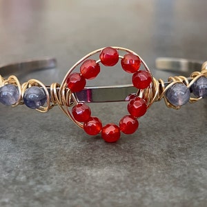 May include: A handcrafted bracelet featuring a silver-tone cuff with gold wire accents. The bracelet is adorned with red and blue-grey beads, and a central design of red beads. A unique piece of jewelry.