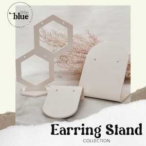 May include: White acrylic earring display stand with three different shapes: a hexagon, a half-circle, and a curved rectangle. The stand is on a white surface. Earring Stand Collection.