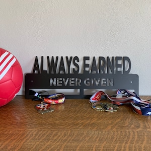 May include: Black metal medal holder with the words "Always Earned Never Given" and three medals hanging from it. A soccer ball is to the left of the holder.