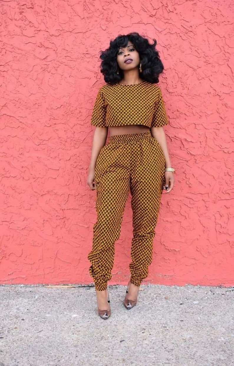 Pu&ograve; includere: Una donna indossa un top corto e pantaloni abbinati con motivo giallo e nero. L'abito &egrave; un completo di due pezzi. &Egrave; in piedi davanti a un muro rosa.
