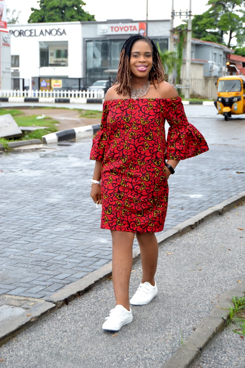 May include: A woman wearing a red and yellow floral off-the-shoulder dress with bell sleeves and white sneakers. She is walking on a sidewalk.