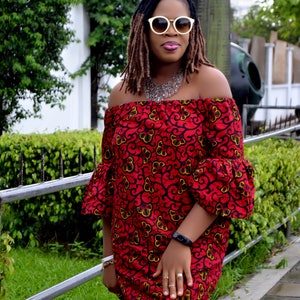 May include: A woman wearing a red off-the-shoulder dress with a floral pattern. The dress has bell sleeves and a fitted bodice. She is wearing white sunglasses and a silver necklace.