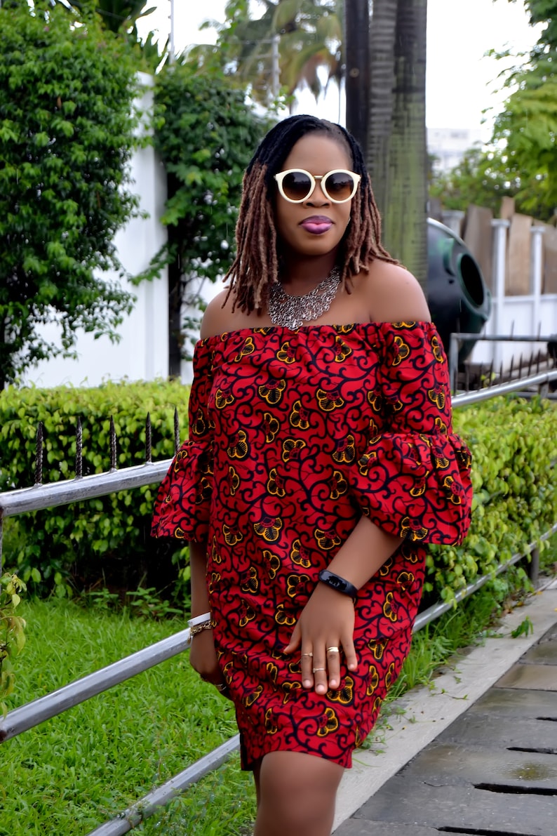 May include: A woman wearing a red off-the-shoulder dress with a floral pattern. The dress has bell sleeves and a fitted bodice. She is wearing white sunglasses and a silver necklace.